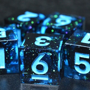 May include: A set of six-sided dice featuring a dark blue and black translucent resin base with embedded green and blue glitter particles. Each die has large, recessed numbers filled with a bright, glowing light blue pigment for high contrast.