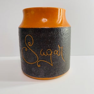 May include: A ceramic sugar jar with a textured black body and a brown rim. The word "sugar" is written in a cursive font on the front of the jar. The jar has a wide opening and a cylindrical shape.