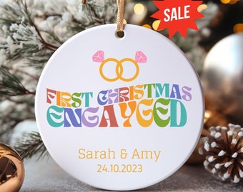 May include: A white ceramic Christmas ornament with gold string. The ornament features the text "FIRST CHRISTMAS ENGAGED" in colorful retro font, with the names "Sarah & Amy" and the date "24.10.2023" below. A red "SALE" tag is visible.