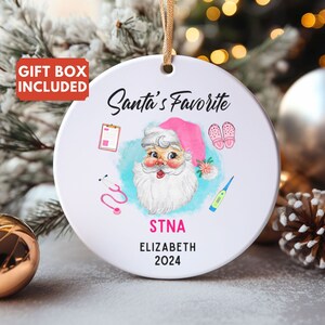Peut inclure: Ornement de Noël rond en céramique blanche avec une ficelle dorée. Il présente une illustration de Père Noël en chapeau rose, avec le texte "Santa's Favorite", "STNA", "Elizabeth 2024" et des icônes médicales.