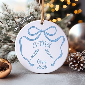 Peut inclure: Ornement de Noël en céramique blanche avec un motif de ruban bleu, le texte "STNA", une seringue, un thermomètre et "Olivia 2025". Cordon doré. Décoration de vacances festive.
