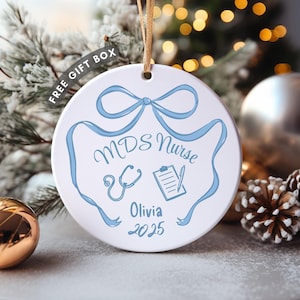 May include: A white ceramic Christmas ornament with a blue ribbon design and the words "MDS Nurse" and "Olivia 2025". The ornament includes a stethoscope and clipboard graphic. A gold string is attached for hanging.