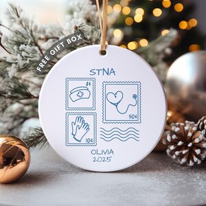 Peut inclure: Ornement de Noël en céramique blanche avec une ficelle dorée. L'ornement présente des illustrations bleues d'un bonnet d'infirmière, d'un stéthoscope, de gants et le texte "STNA" et "OLIVIA 2025".