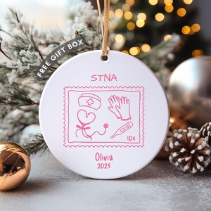 Peut inclure: Ornement de Noël en céramique blanche avec une ficelle dorée. Il présente un motif de timbre rose avec des symboles médicaux et le texte "STNA", "Olivia" et "2025". Un ornement doré et argenté est en arrière-plan.