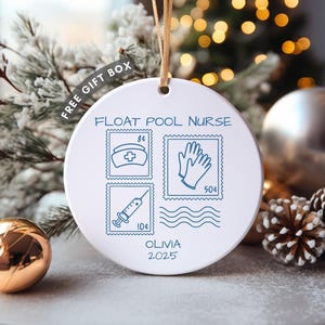 May include: White round Christmas ornament with the text "FLOAT POOL NURSE" and blue illustrations of a nurse's hat, gloves, and a syringe. The ornament also includes the name "OLIVIA" and the year "2025". A gold string is attached for hanging.