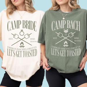 May include: Two t-shirts, one cream and one olive green, with the text "CAMP BRIDE" and "CAMP BACH" respectively. Both shirts feature a graphic with crossed marshmallow roasting sticks, a ring, and the text "LET'S GET TOASTED". The shirts are for a bachelorette party.