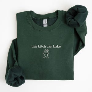 Może przedstawiać: Leśnozielona bluza z haftowanym napisem "this bitch can bake" i piernikowym ludzikiem w czapce Świętego Mikołaja. Top z okrągłym dekoltem i długimi rękawami wykonany jest z miękkiego materiału.