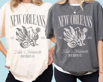 Puede incluir: Dos camisetas, una marfil y otra pimienta, cada una con un gráfico de Nueva Orleans. El diseño incluye una máscara, un cóctel y el texto "New Orleans", "Kate's Nolarette" y "Bourbon St."