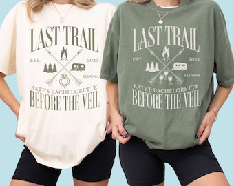 May include: Two t-shirts, one cream and one olive green, with the text "LAST TRAIL" above a graphic of crossed arrows, a campfire, and a camper. The text "KATE'S BACHELORETTE BEFORE THE VEIL" is below the graphic.