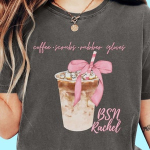 May include: Dark gray t-shirt featuring a watercolor illustration of an iced coffee drink with a pink bow and straw. The shirt has the text "coffee scrubs rubber gloves" and "BSN Rachel" in pink script.