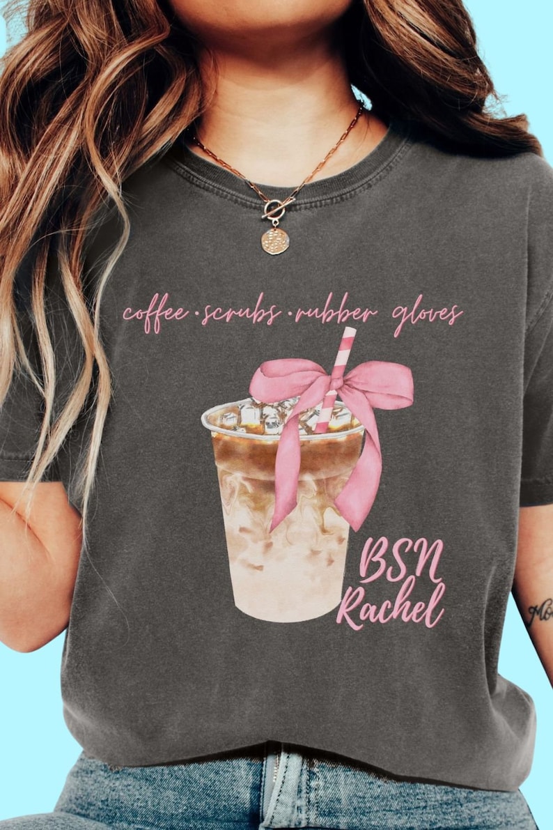 May include: Dark gray t-shirt featuring a watercolor illustration of an iced coffee drink with a pink bow and straw. The shirt has the text "coffee scrubs rubber gloves" and "BSN Rachel" in pink script.