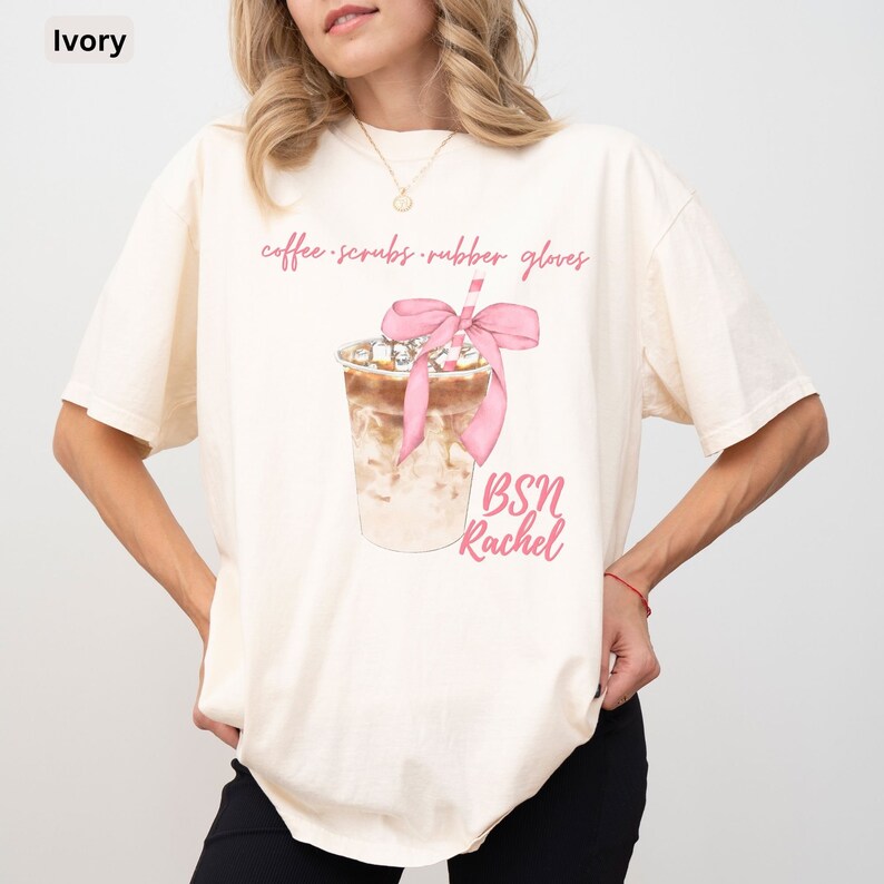 May include: Ivory-colored t-shirt featuring a watercolor illustration of an iced coffee drink with a pink bow and straw. The text "coffee scrubs rubber gloves" is above the drink, and "BSN Rachel" is below it.