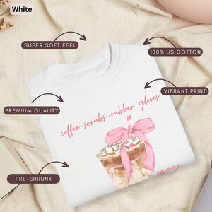 May include: White t-shirt with a watercolor illustration of an iced coffee drink with a pink bow and straw. The text "coffee scrubs rubber gloves" is written in pink script. Features include 100% US cotton, pre-shrunk, premium quality, and a super soft feel.