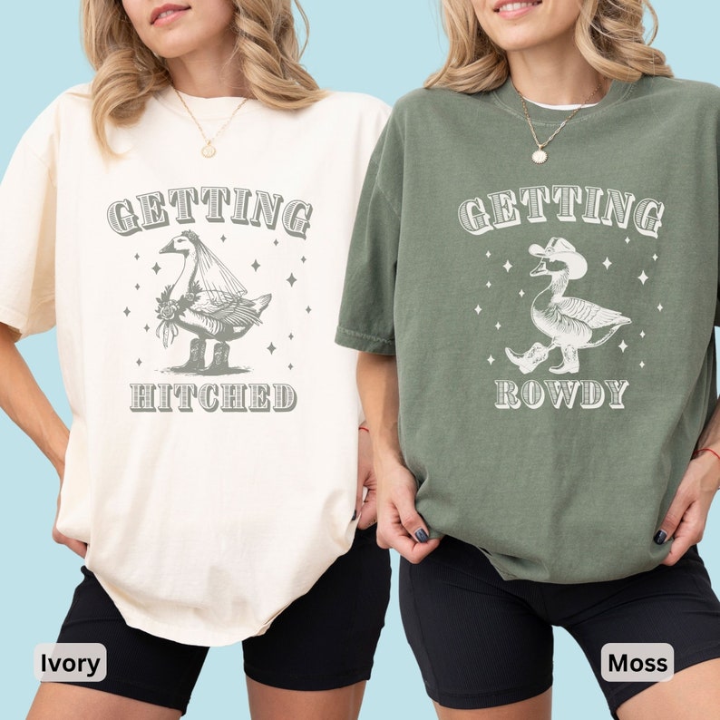 Puede incluir: Dos camisetas, una marfil y otra verde musgo, cada una con un gr&aacute;fico de un ganso. La camiseta marfil dice "Getting Hitched" y la camiseta verde musgo dice "Getting Rowdy". Ambas camisetas tienen un aspecto vintage y desgastado.