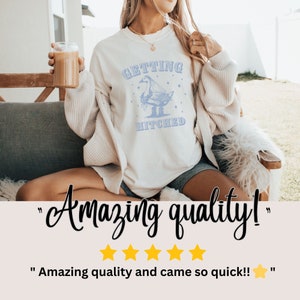 Puede incluir: Camiseta color crema con un gr&aacute;fico azul claro de un ganso y las palabras "GETTING HITCHED." La camiseta la lleva una persona. La imagen tambi&eacute;n incluye el texto "Amazing quality!" y cinco estrellas doradas.