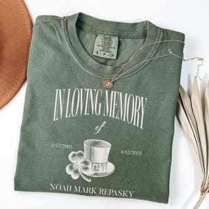 May include: An olive green t-shirt with the text "IN LOVING MEMORY of" and dates 3/17/2001 and 4/12/2025. A graphic of a top hat and shamrock is below the text. A gold necklace with a pendant rests on the shirt.