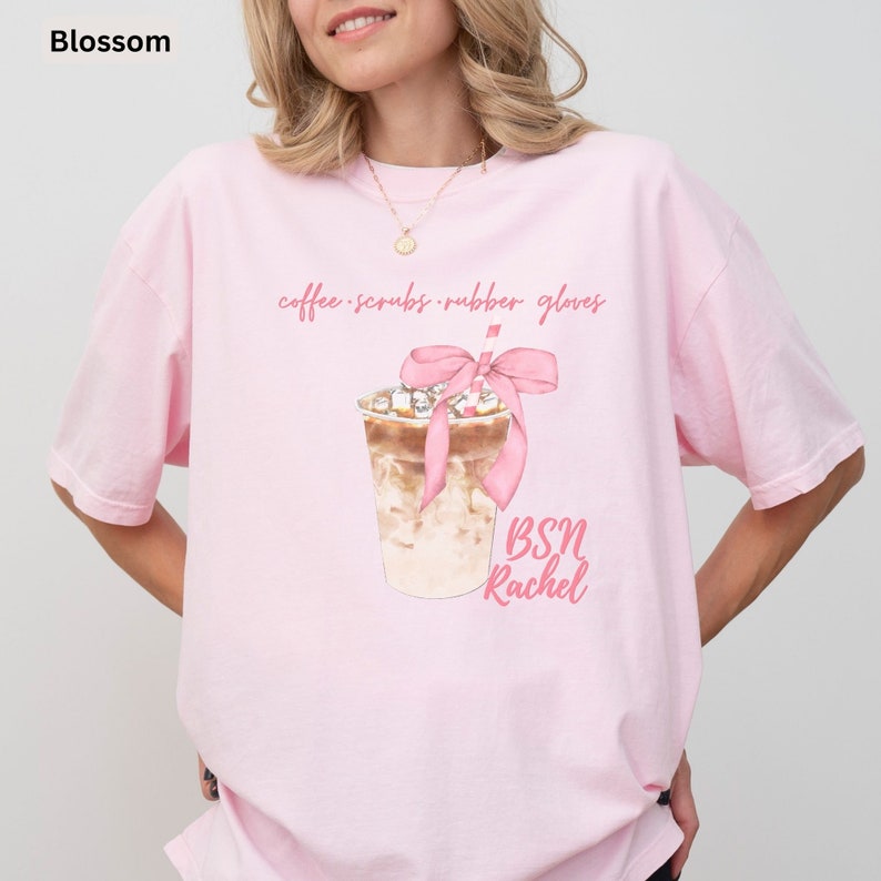 May include: Light pink t-shirt with the word "Blossom" in the upper left corner. The shirt features a watercolor illustration of an iced coffee drink with a pink bow and the text "coffee scrubs rubber gloves" and "BSN Rachel".