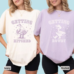 Puede incluir: Dos camisetas, marfil y orqu&iacute;dea, con los textos "GETTING HITCHED" y "GETTING ROWDY". Cada camiseta presenta un gr&aacute;fico caprichoso de un ganso. La camiseta marfil tiene un ganso con velo, mientras que la camiseta orqu&iacute;dea tiene un ganso con sombrero de vaquero.