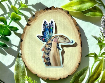 May include: A blue and purple butterfly sticker with glitter accents is holding a small brown and white lizard with its wings. The sticker is on a wooden background.