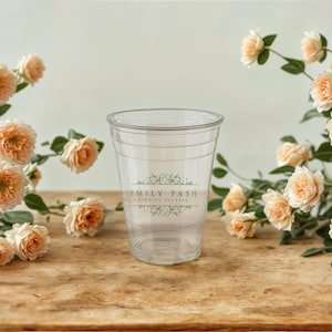 May include: A clear plastic cup with a white label that says "Emily Pash Wedding Florist". The cup is surrounded by peach-colored roses.