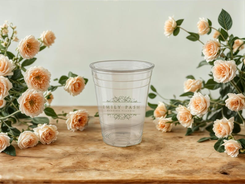 May include: A clear plastic cup with a white label that says "Emily Pash Wedding Florist". The cup is surrounded by peach-colored roses.