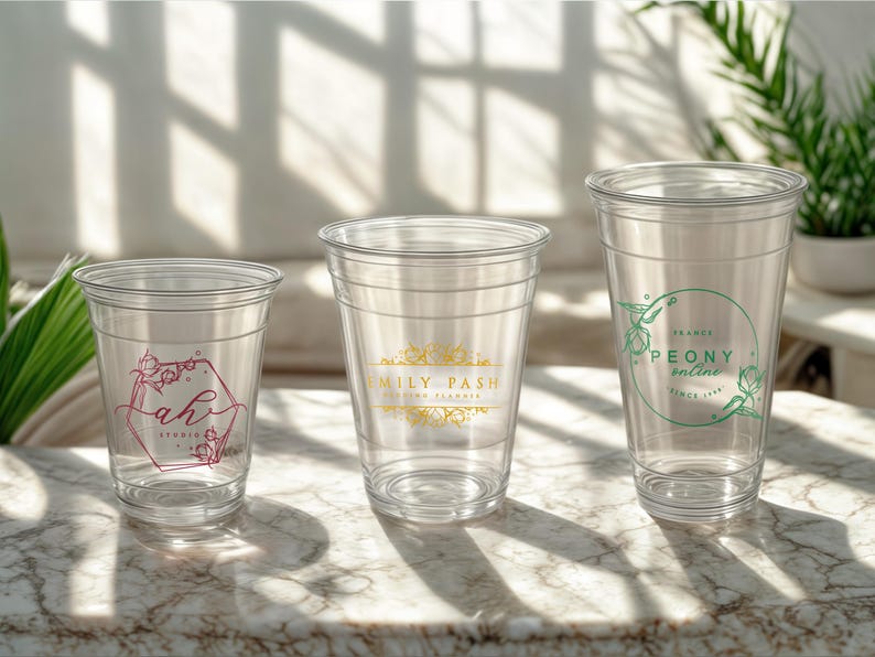 May include: Three clear plastic cups with different floral designs. The cup on the left has a pink floral design with the text "ah! studio". The middle cup has a gold floral design with the text "Family Pash". The cup on the right has a green floral design with the text "PEONY online since 1962".