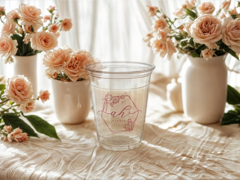 May include: A clear plastic cup with a pink and white floral design and the text "ah! studio" on the side. The cup is sitting on a white tablecloth with a bouquet of peach roses in the background.