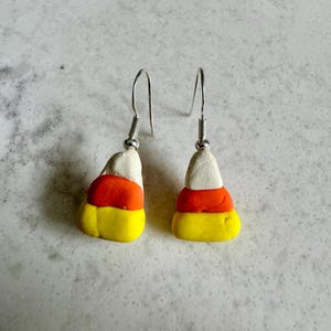 May include: A pair of dangle earrings shaped like candy corn. The earrings are made of polymer clay and have a yellow, orange, and white color scheme.