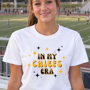 May include: A white short-sleeved t-shirt featuring the text "IN MY CHIEFS ERA" in a bold, yellow, retro-style font with black outlines. The design is surrounded by small, four-pointed black and yellow star icons scattered around the text.
