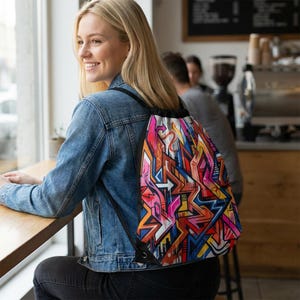 May include: A drawstring backpack with a vibrant graffiti design. The bag features a black drawstring closure and straps. The graffiti art includes a mix of bright colors, including pink, orange, blue, and yellow, with white accents. The bag is worn over the shoulder.