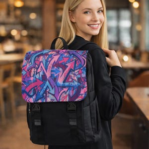 Puede incluir: Mochila negra con solapa con un colorido diseño de graffiti de flechas en tonos rosa, morado y azul. La mochila tiene un asa superior y correas ajustables.