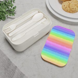 May include: A white rectangular lunch box with a fork, spoon, and knife inside. A colorful rainbow-striped coaster is next to the lunch box. The coaster has a cork base and a watercolor design.