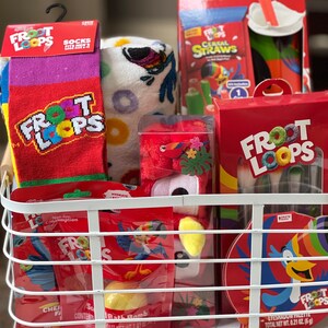 May include: A white wire basket filled with Froot Loops-themed merchandise. Items include socks, a towel, bath bombs, cereal straws, and an eyeshadow palette. The socks are red with the Froot Loops logo. The towel has a colorful design.