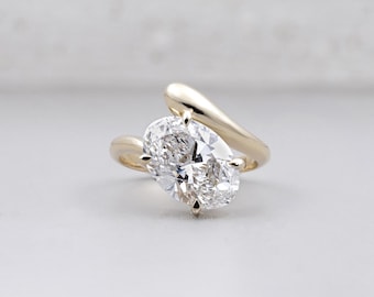 Può includere: Un anello d'oro con un grande diamante taglio ovale. Il diamante è il punto focale, incastonato con quattro griffe. La fascia ha un design fluido e contemporaneo. L'anello è posto su uno sfondo neutro.