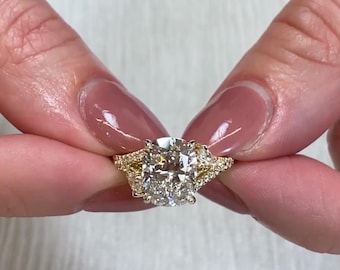 Könnte beinhalten: Ein goldener Ring mit einem großen, funkelnden Diamanten im Kissenschliff. Der Ring hat kleinere Diamanten auf beiden Seiten des Hauptsteins und entlang des Bandes, wodurch ein schillerndes Spiel aus Licht und Brillanz entsteht.