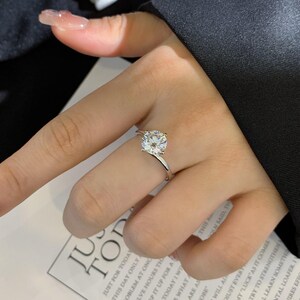 May include: A silver ring with a large, round, clear gemstone. The ring's band curves gracefully towards the stone. The ring is on a finger. The background includes a portion of a black garment and a printed page.