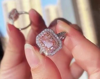 May include: A close-up of a pink gemstone ring with a halo of smaller, clear stones. The ring has a rectangular center stone and a silver band. Another ring is visible in the background, out of focus.