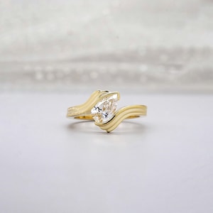 Könnte beinhalten: Ein goldener Ring mit einem birnenförmigen Diamanten. Der Ring hat ein einzigartiges, fließendes Design mit einem wellenartigen Band. Der Diamant ist in einer Zargenfassung gefasst. Der Ring wird auf einer weißen Oberfläche präsentiert.