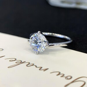May include: A silver engagement ring with a large, round, clear gemstone. The ring has a simple band and four prongs holding the gemstone. The ring is set against a white and black background.