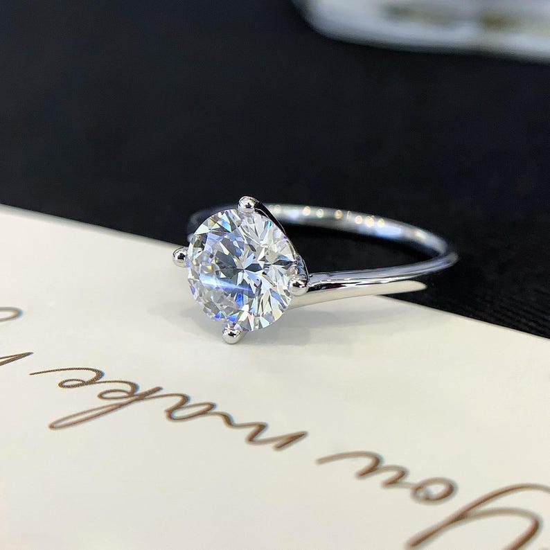 May include: A silver engagement ring with a large, round, clear gemstone. The ring has a simple band and four prongs holding the gemstone. The ring is set against a white and black background.