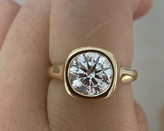 Puede incluir: Un anillo de oro con una piedra preciosa transparente de talla brillante redonda engastada en un bisel cuadrado liso. La banda es de oro pulido y la piedra central muestra múltiples facetas que reflejan la luz, creando un aspecto brillante.
