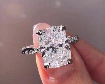 May include: A silver ring featuring a large, cushion-cut diamond. The diamond is set in a prong setting and has a halo of smaller diamonds around it. The band is also set with smaller diamonds.