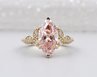May include: A marquise-cut engagement ring featuring a large, pale pink gemstone. The gold band has detailed leaf and dot patterns, with small accent stones. The setting is ornate and elegant, with a vintage style.