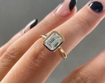 Könnte beinhalten: Ein goldener Ring mit einem Diamanten im Smaragdschliff. Der Diamant ist in einer Lünettenfassung gefasst, und der Ring hat ein schlichtes, elegantes Design. Der Ring wird an einem Finger mit neutraler Maniküre gezeigt.