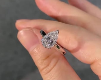 Puede incluir: Un anillo de metal plateado con una gema transparente facetada en forma de pera engastada en garras. La banda es lisa y pulida, mostrando un acabado metálico reflectante que complementa el corte brillante de la piedra central.