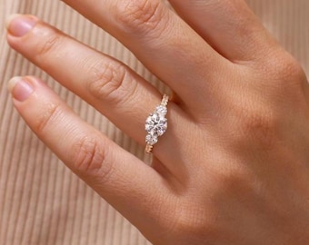 May include: A three-stone ring featuring a large round-cut center diamond flanked by two smaller round-cut diamonds on a thin gold band. The stones are set in a prong setting, creating a classic look with clear, sparkling white gemstones.