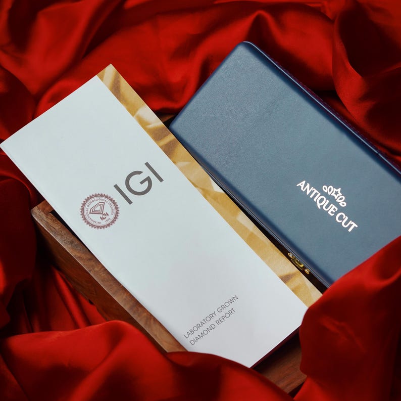 May include: A white IGI Laboratory Grown Diamond Report and a blue box with "ANTIQUE CUT" printed on it. The report is in a wooden box, set against a red satin background.