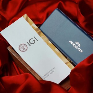 May include: A white IGI Laboratory Grown Diamond Report rests in a wooden box next to a blue box with "ANTIQUE CUT" printed on it. The items are set against a backdrop of red fabric.