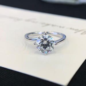 May include: A silver engagement ring with a large, round, clear gemstone. The gemstone is held in place by four prongs. The ring is set on a white surface.