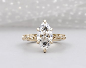 May include: A marquise-cut diamond engagement ring with a gold band. The band features delicate leaf and dot engravings. The diamond is set with six prongs and sparkles brightly. A classic and elegant piece of jewelry.